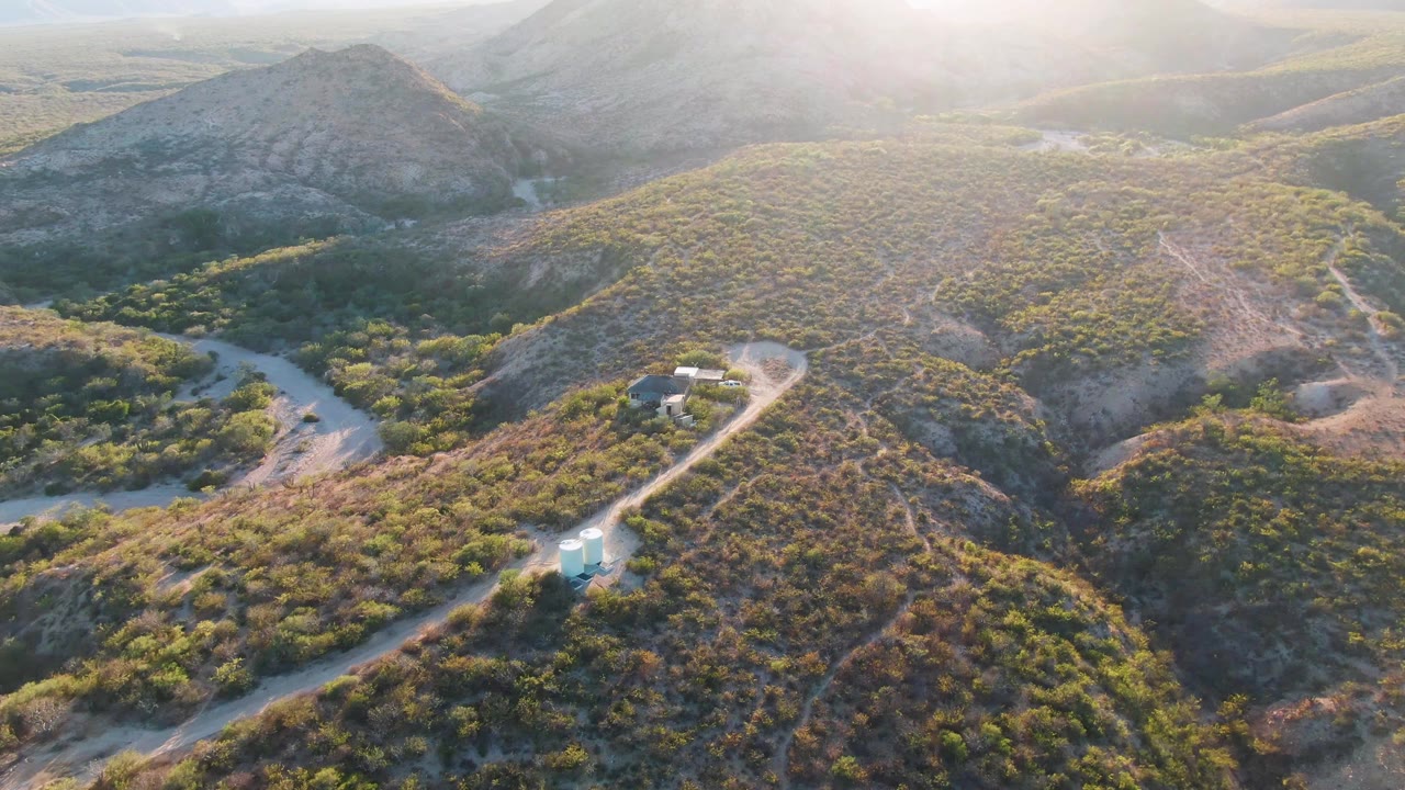 Aerial frame from Cabo Pulmo, Baja California Sur, Mexico, showing a light-colored vehicle near the lower road, scrubby hills, a small roadside structure, warm haze, and no visible people.