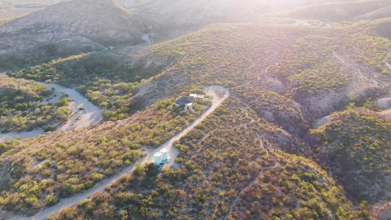 High oblique aerial frame from Cabo Pulmo, Baja California Sur, Mexico, showing the vehicle on the pale dirt road below scrub-covered slopes, a small structure nearby, and no visible people.