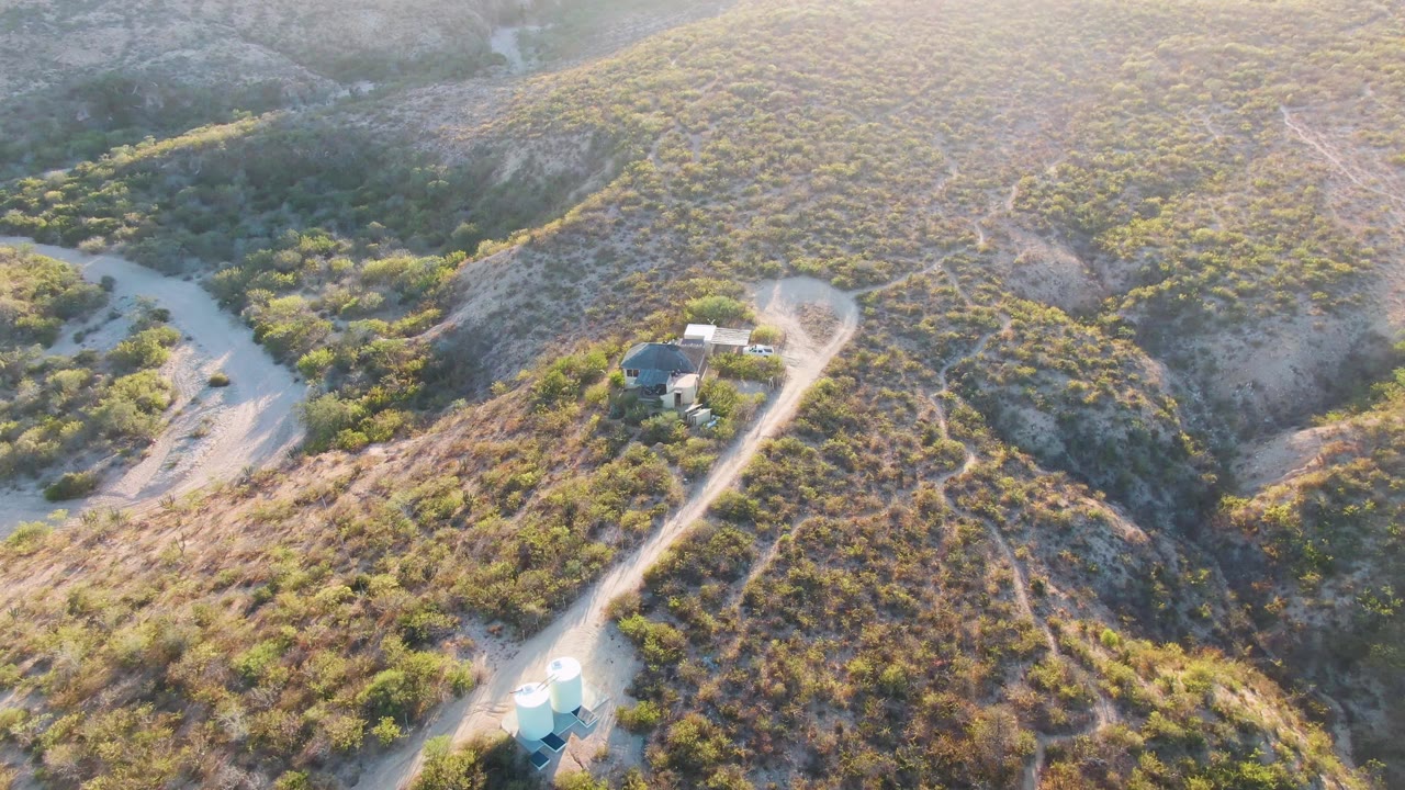 Aerial frame from Cabo Pulmo, Baja California Sur showing a light van near the lower road bend, a small structure on the hillside, dry brush, tan soil, and no visible people.