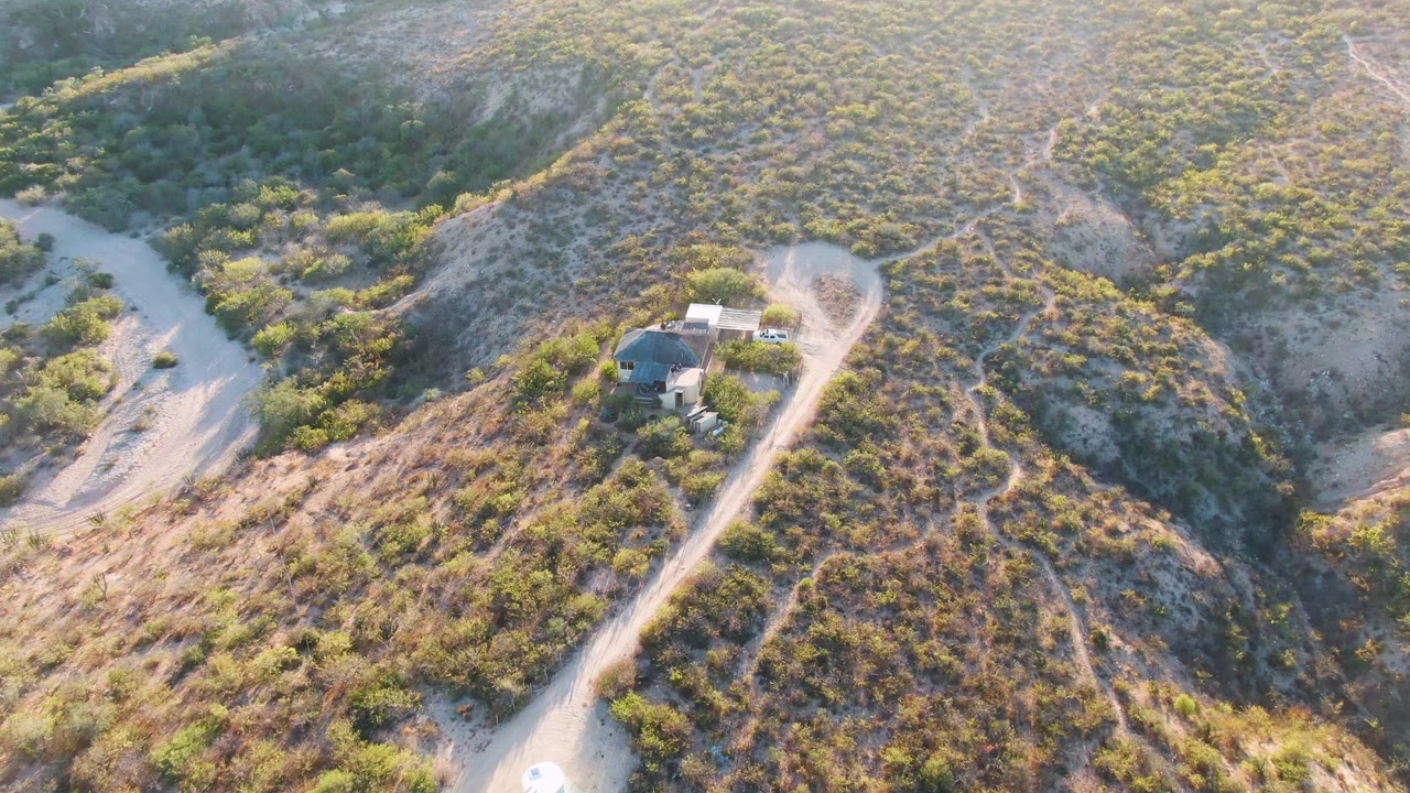 Aerial frame from Cabo Pulmo, Baja California Sur showing the winding road, a small structure near the center-right hillside, scrub vegetation, pale exposed soil, and no visible people.