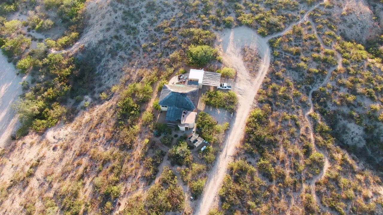 Aerial frame from Cabo Pulmo, Baja California Sur showing the house higher in the composition, branching dirt tracks, dry brown ground, green shrubs, and no visible people.