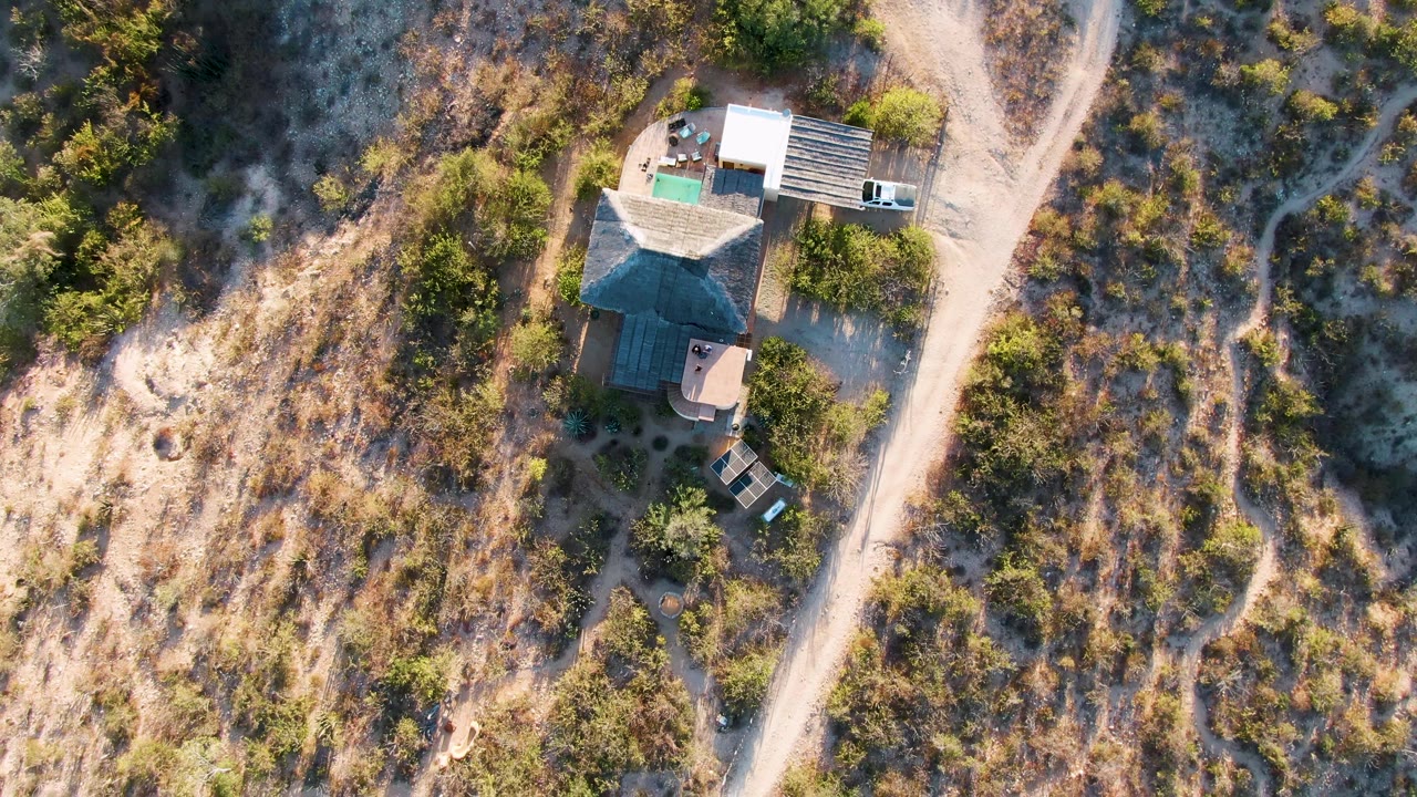 One second in, the aerial view holds over the Cabo Pulmo house as the dirt road passes along the right side of frame, with scrub vegetation, dry hillside ground, and no visible people.