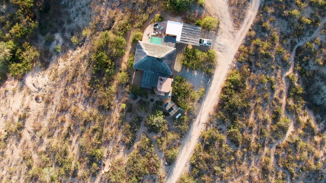 At 0:02, the house remains near the left of the road in Cabo Pulmo, with the white truck at the road edge, a parked car by the building, brushy dry ground, and no visible people.