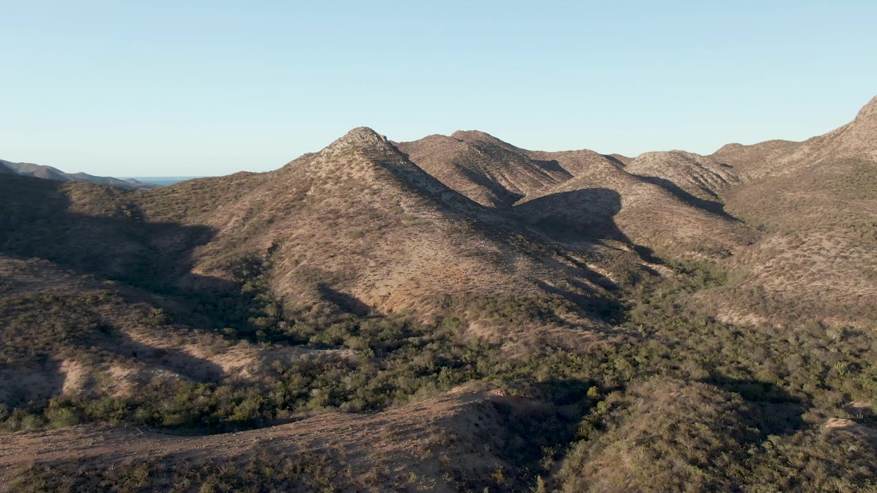 Opening aerial view of dry ridges near Cabo Pulmo, Baja California Sur, with a pointed central hill, scattered green vegetation in the valley, and no visible people.