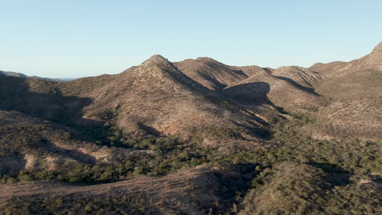 Early frame from the Cabo Pulmo mountain aerial showing a brown foreground slope, a shadowed valley with scattered trees, layered hills, and no visible people.