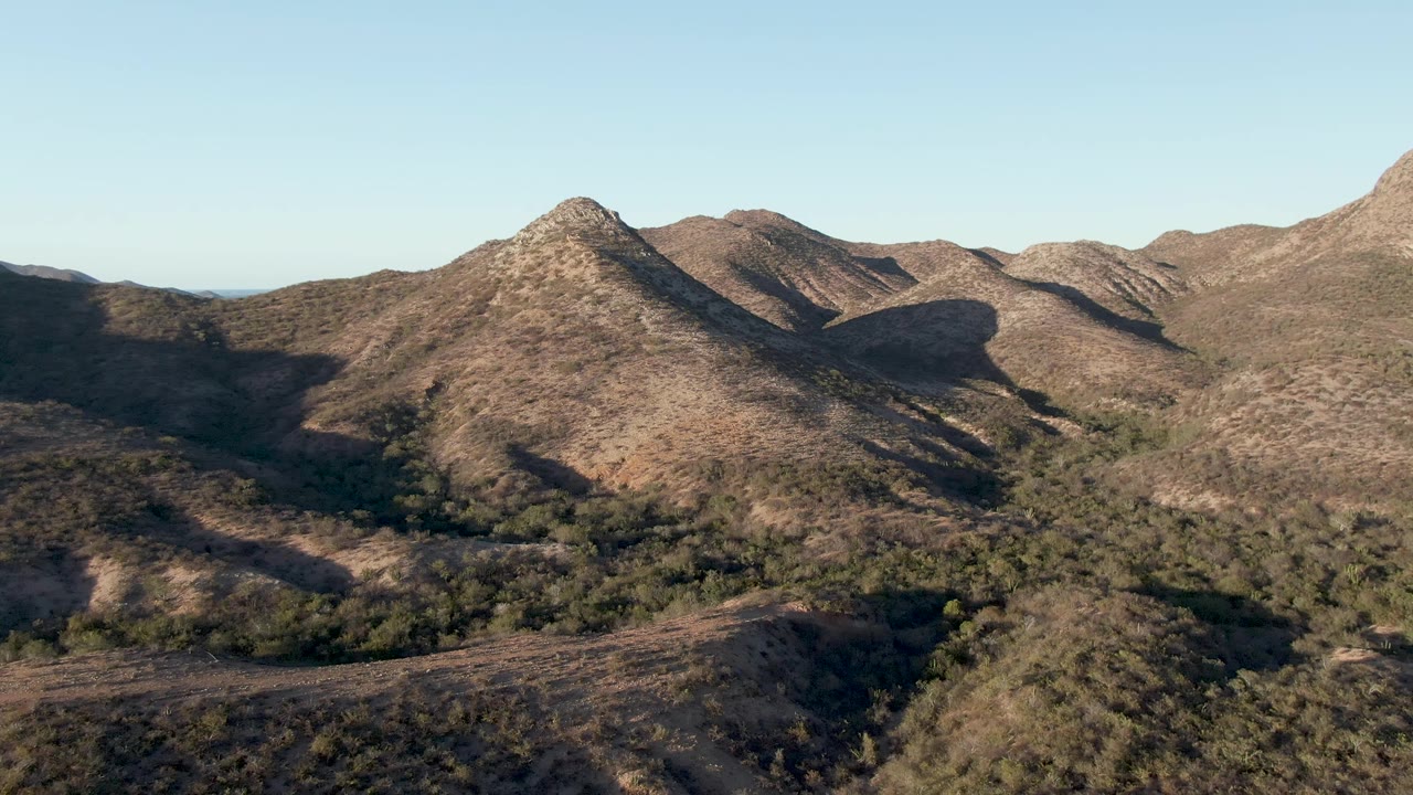 Aerial frame over Cabo Pulmo terrain with the pointed ridge centered, dry brown hills extending behind it, patchy green trees below, and no visible people.