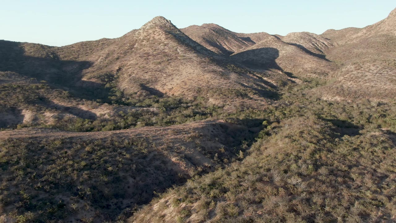 Aerial frame over Cabo Pulmo, Baja California Sur, showing dry brown ridges, scattered green vegetation, a pale sky, and no visible people at the start of the clip.