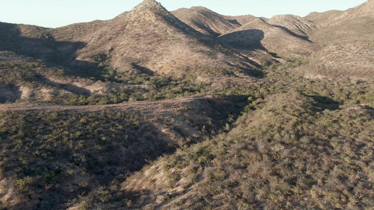 Aerial view in Cabo Pulmo, Baja California Sur, with a closer foreground slope, layered dry ridges, scattered shrubs, and no visible people.