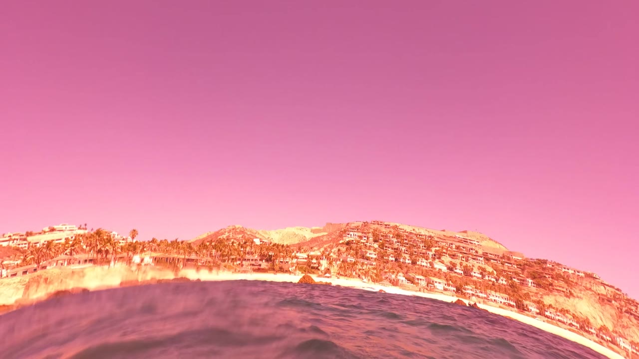 The camera is at the ocean surface looking toward the San Jose del Cabo coastline, with hills, buildings, waves, and a pink color cast, with no visible people.