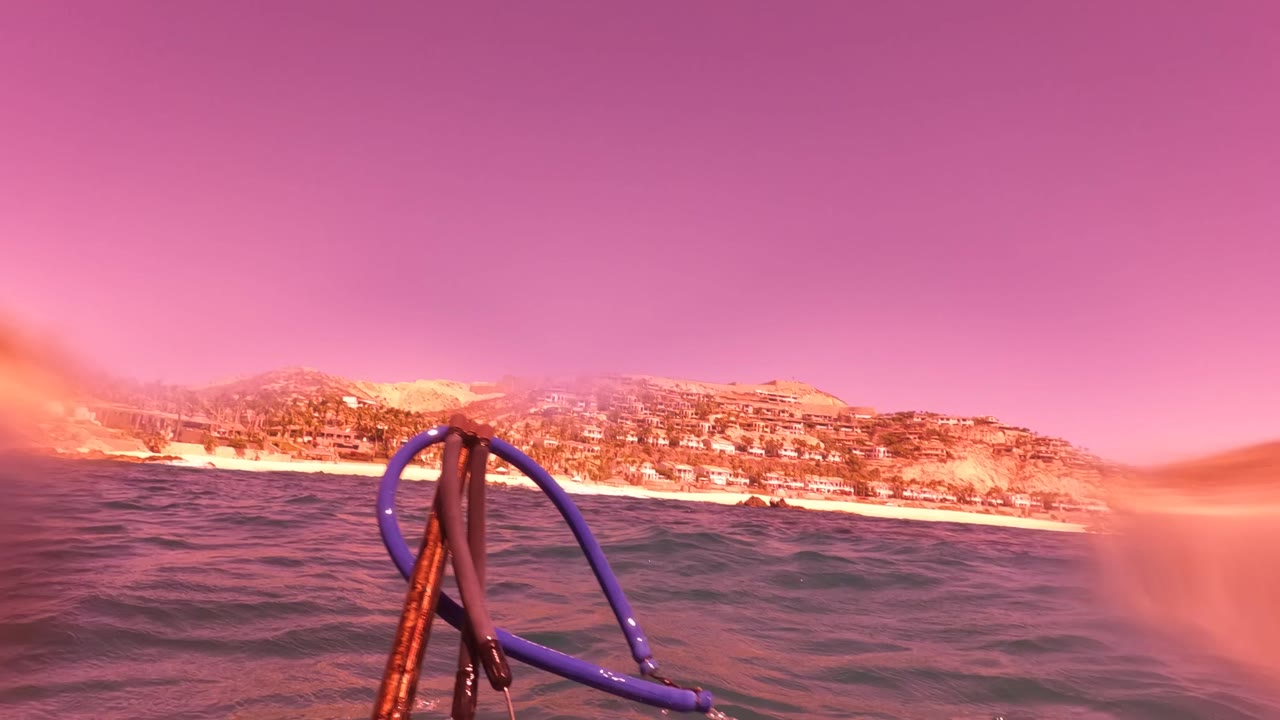 A blue rope and darker rigging sit in the foreground above choppy water, with San Jose del Cabo hills and shoreline buildings in the background and no visible people.