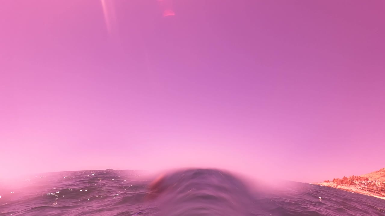 At 0:00 in San Jose del Cabo, a pink-purple ocean surface fills the frame with a small wave crest in front, coastline at right, and no clearly visible person.