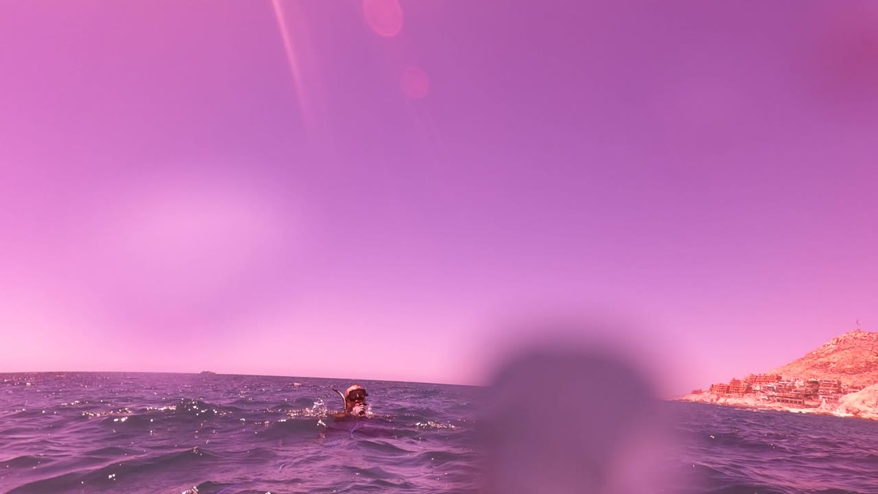 At 0:02 in San Jose del Cabo, the swimmer is farther right near the horizon, partly framed by a large soft foreground droplet and purple-toned sea.
