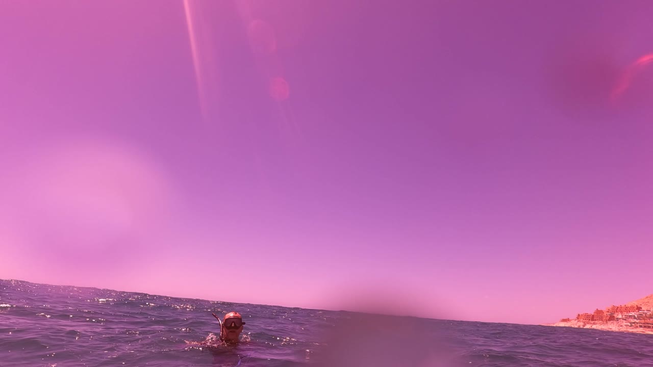 A swimmer is centered low in the San Jose Del Cabo ocean, with choppy dark water, a pink sky, and rocky shoreline visible along the right edge.