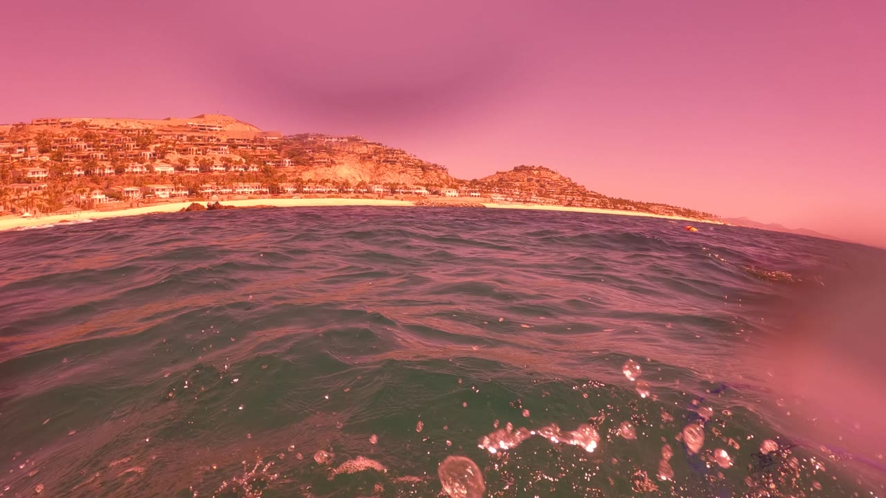 Surface-level view of choppy ocean water near the sandy San Jose del Cabo coastline, with hillside buildings in the background and no visible people.