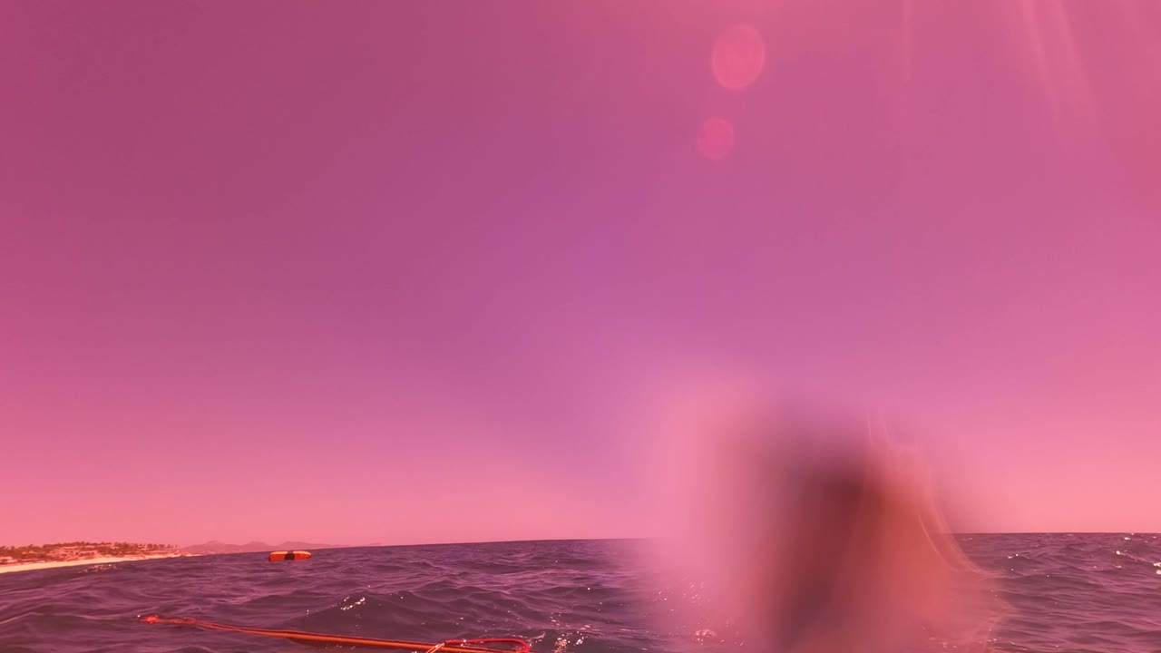 Water-level view of the ocean in San Jose Del Cabo, Mexico, with a pink sky, distant shoreline, orange float line, and blurred water droplets near the lens.