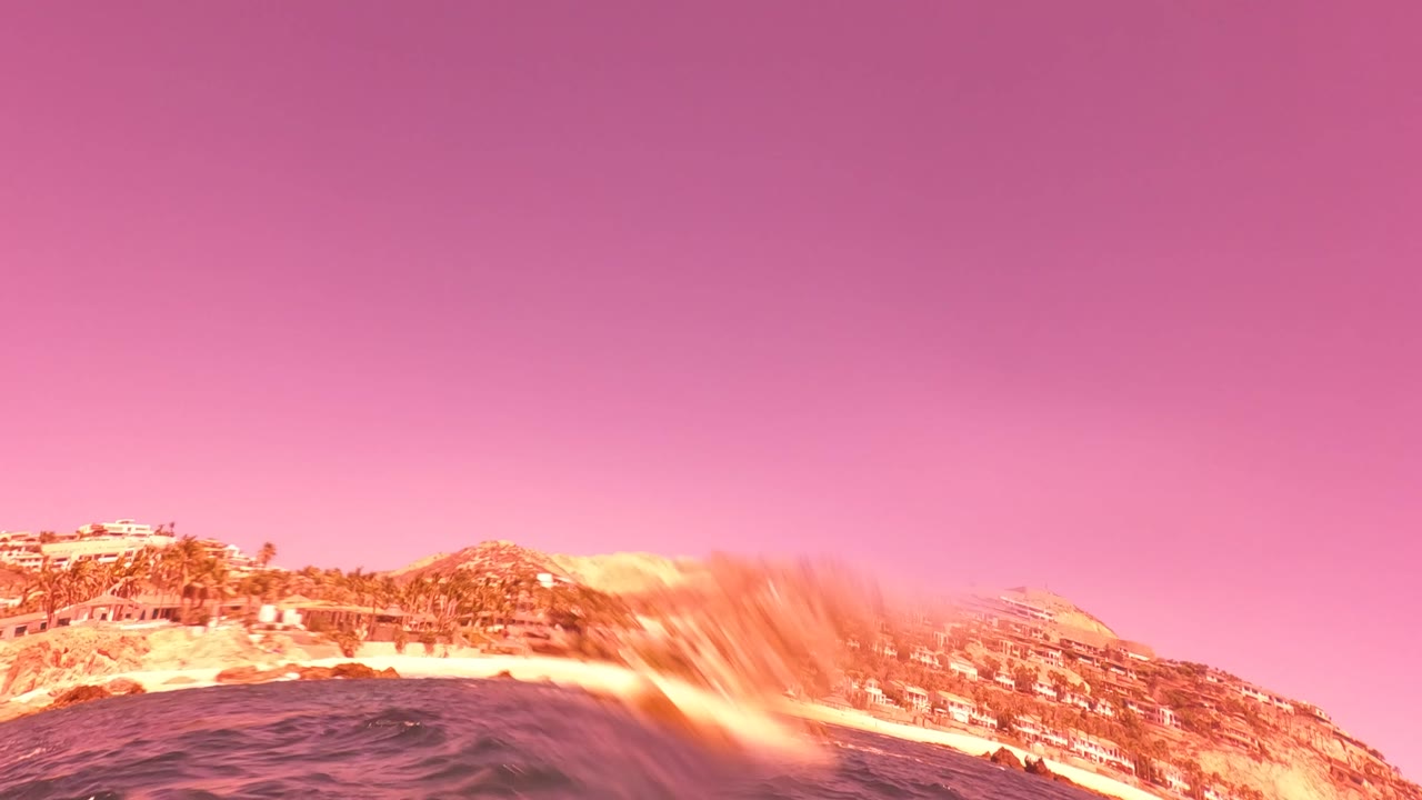 A waterline view in San Jose del Cabo reveals a hillside coastline, buildings in the distance, dark ocean surface, and pink-purple sky, with no visible people.