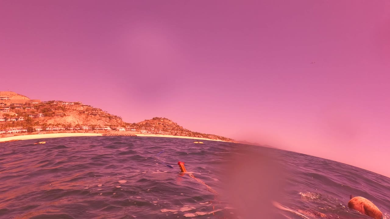 Wave-level frame in San Jose Del Cabo with the horizon tilted slightly by the water, pink sky above, rocky coast in the background, and no visible people.