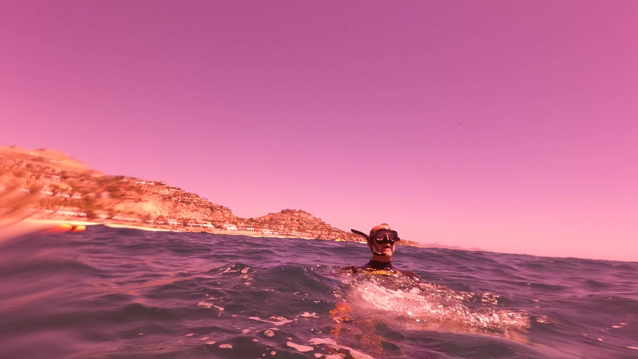 A snorkeler faces the camera from the ocean surface in San Jose del Cabo, with a low wave rising across the frame and a sloping coastal hill behind.