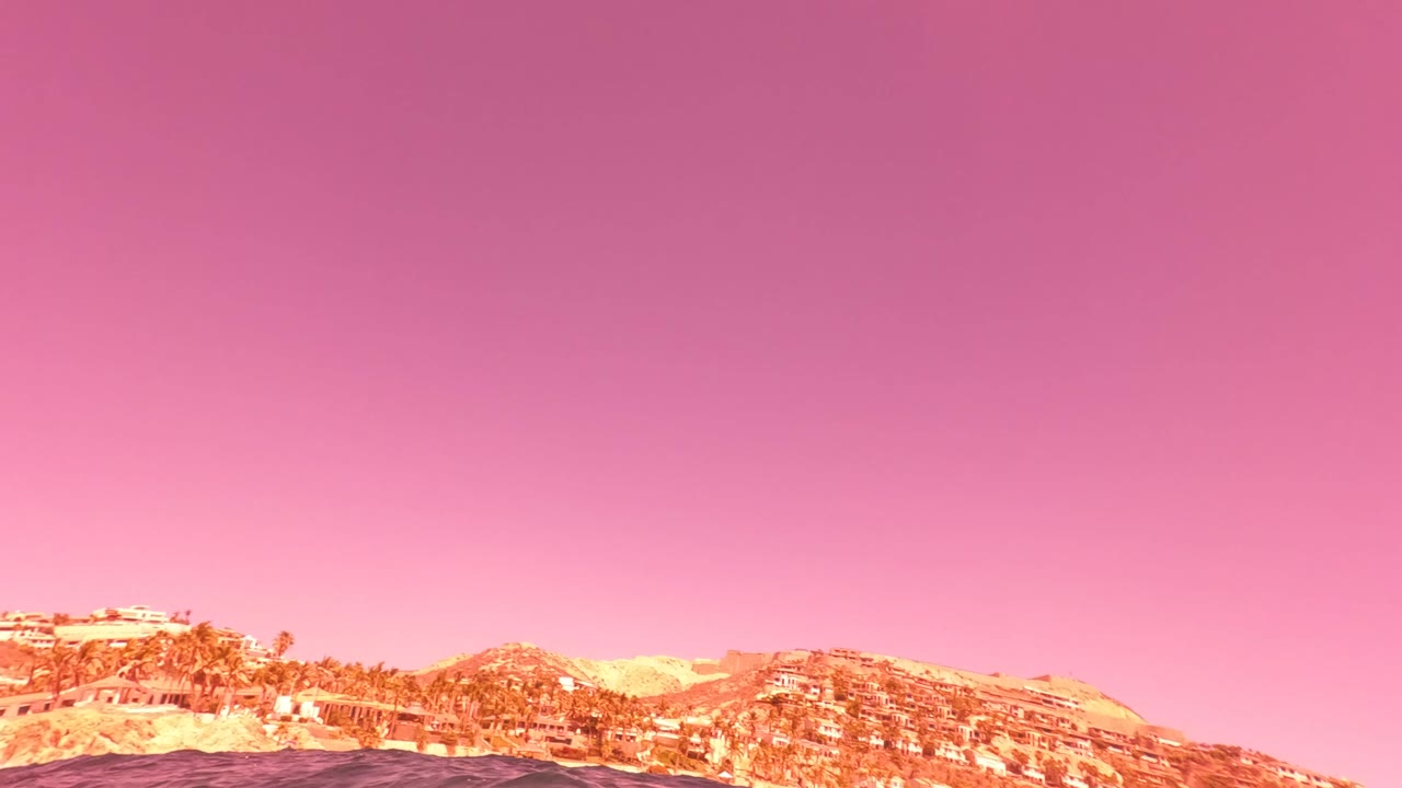 Surface-level view in San Jose del Cabo with dark ocean in the foreground, hills and shoreline buildings in the distance, a purple-pink sky above, and no visible people.