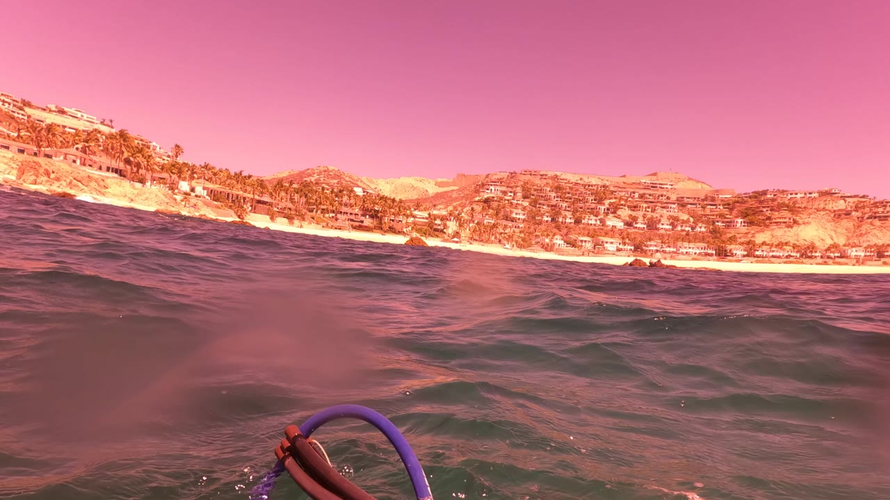 Surface frame in San Jose del Cabo with rippled ocean foreground, hillside coast in the distance, part of a purple snorkel float at the lower right, and no visible people.