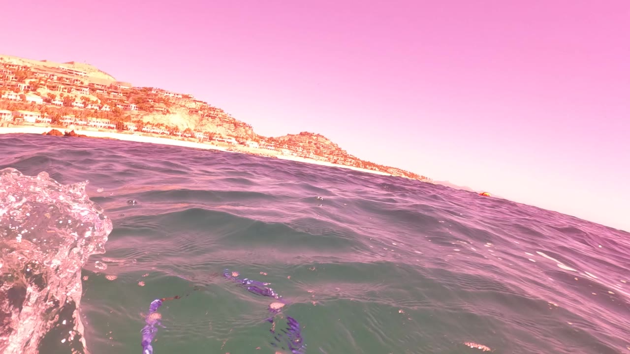 A waterline view shows choppy ocean, a pink-tinted sky, shoreline, and low hills in San Jose del Cabo, with no visible people.