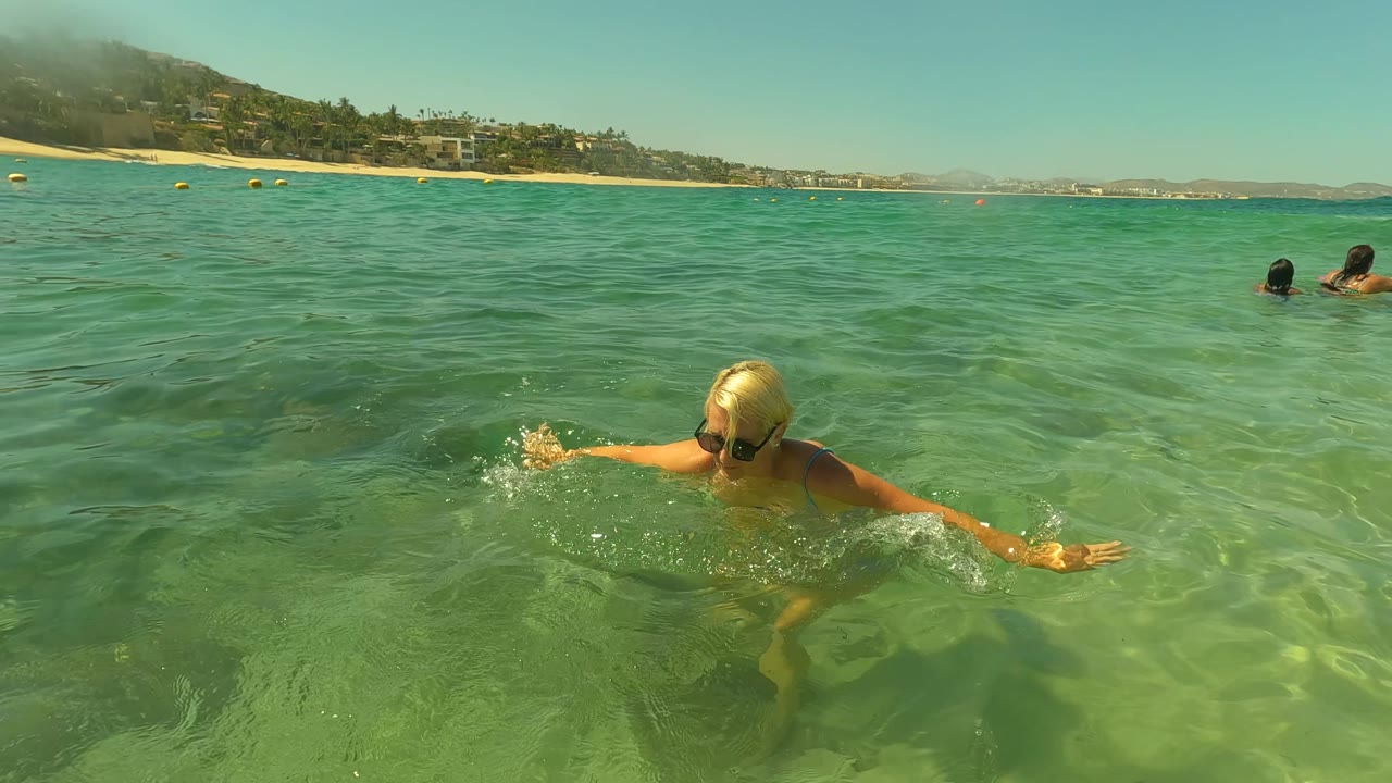 Blonde woman in sunglasses swims at water level in shallow green ocean water in San Jose del Cabo, with beach, hillside buildings, and distant buoys behind her.
