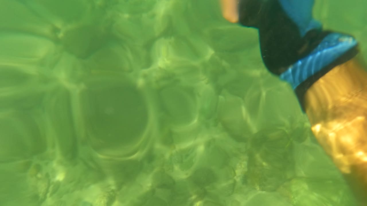 San Jose Del Cabo underwater frame with rippled green water, rocks beneath the surface, faint fish shapes, and the edge of a blue-and-black glove appearing along the upper right border.