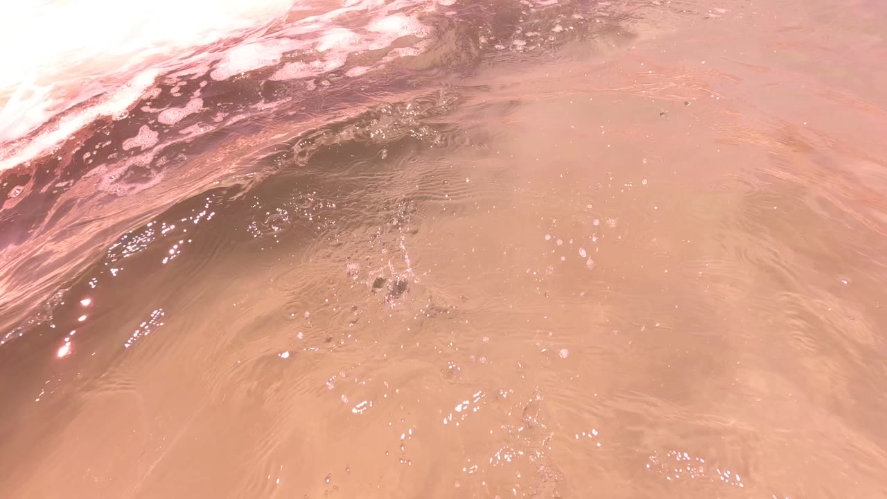 The lens sits at the waterline in San Jose Del Cabo, showing pinkish foam and brown sandy water with no visible people.