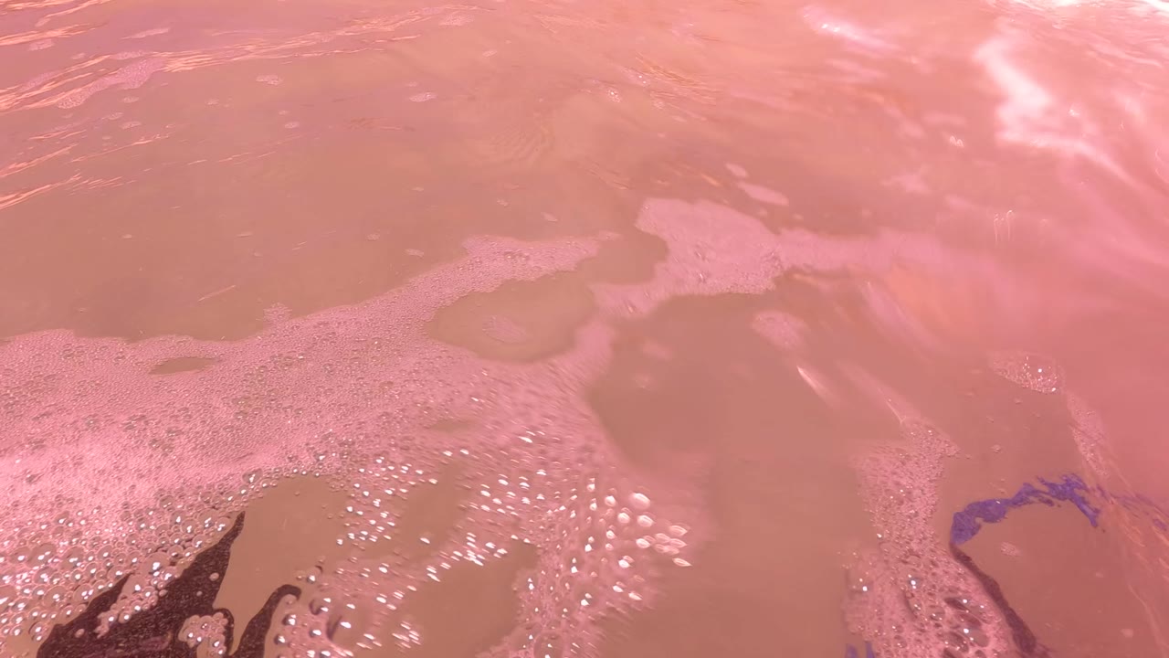 Pink water surface in San Jose del Cabo with foam patches, bright glare, and a small blue curved reflection near the lower center.