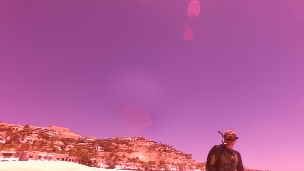 A person stands near the San Jose Del Cabo beach shoreline under a purple sky, with hillside buildings and sand visible behind them.