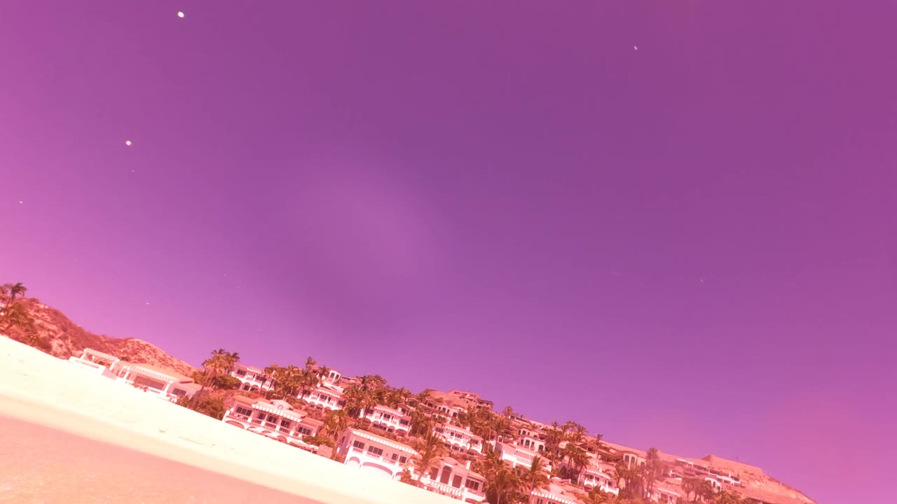 San Jose Del Cabo beach appears at a diagonal with bright sand, beachfront buildings, palms, hills, and purple evening sky, with no visible people.