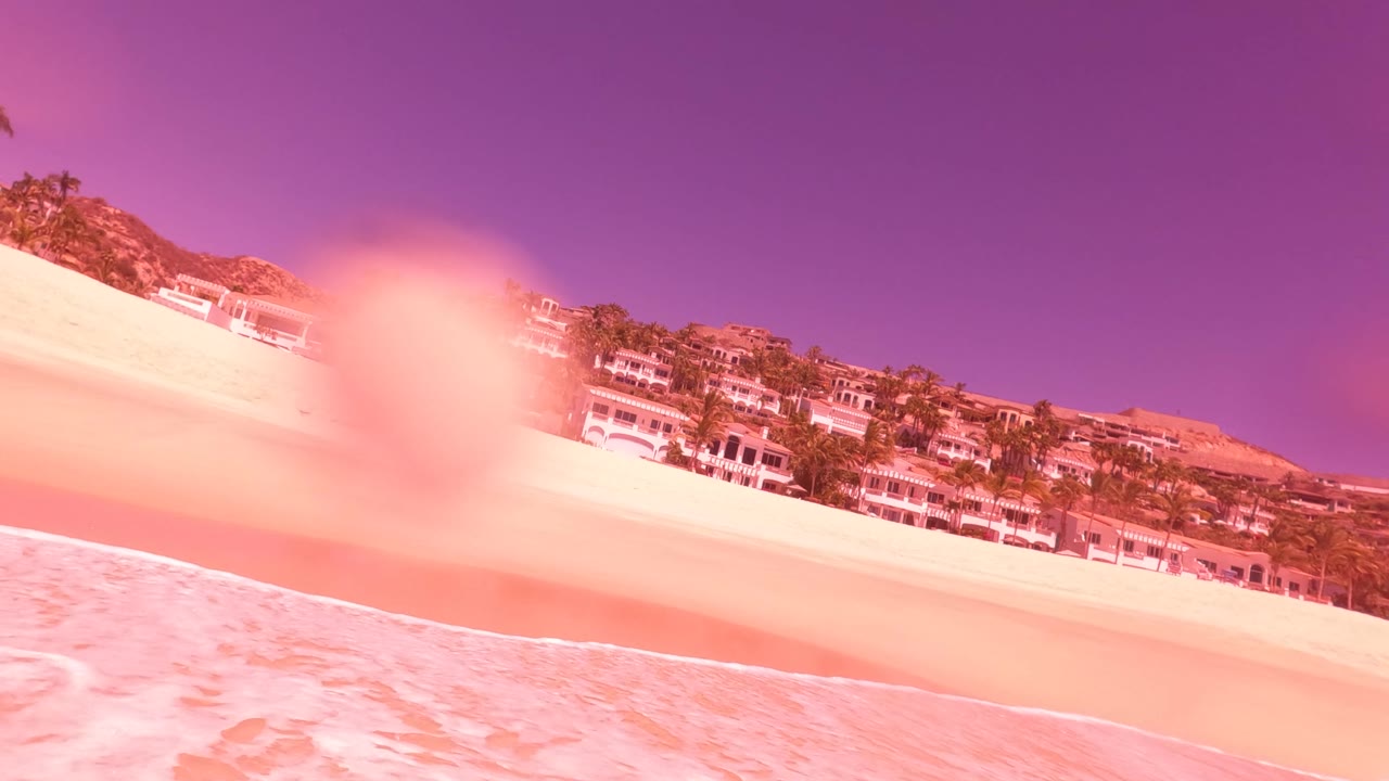San Jose del Cabo shoreline with shallow surf, a man partly visible at the right edge holding gear and tow rope, hillside buildings, palm trees, and strong pink-purple tint.
