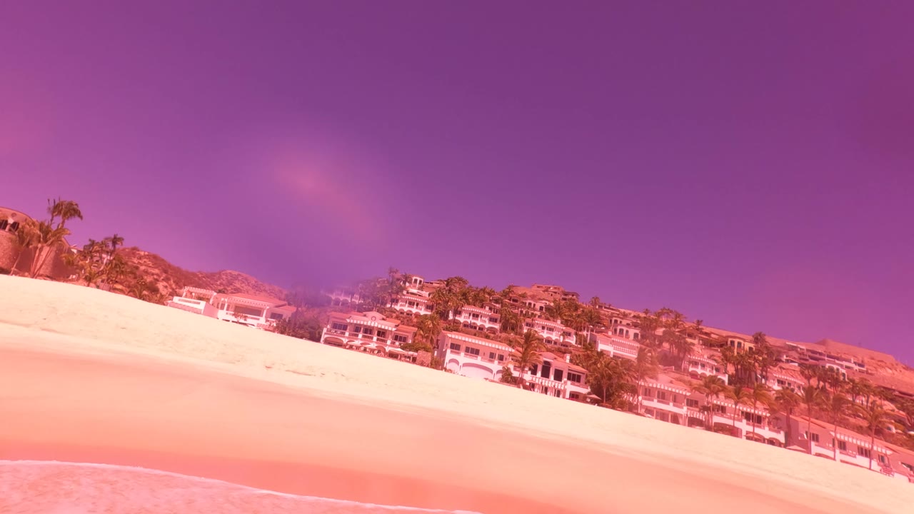 Tilted San Jose del Cabo beach frame showing pale sand, a narrow surf line, hillside buildings, palm trees, and pink-purple color treatment with no visible people.