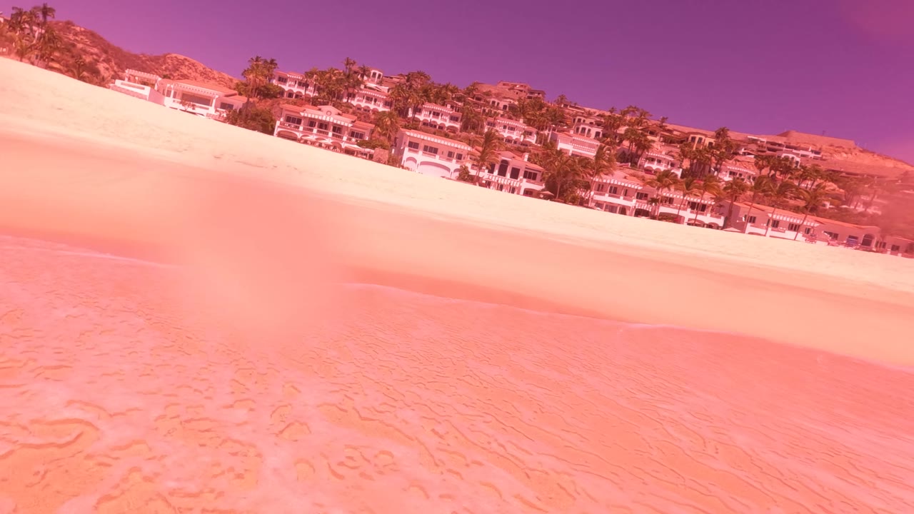 Close shoreline angle with sand, surf edge, hillside buildings, palm trees, and tinted sky.