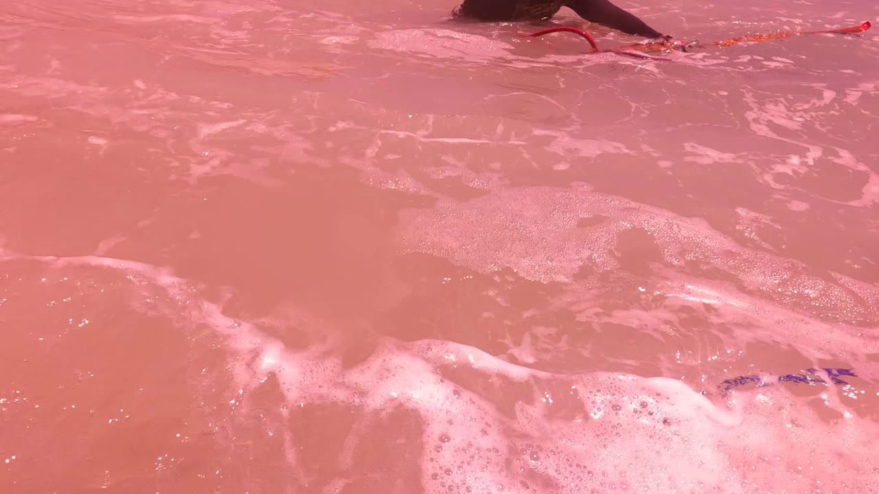 Pink-tinted surf in San Jose del Cabo fills the frame with white foam and a cropped swimmer at the top edge, with no face visible.