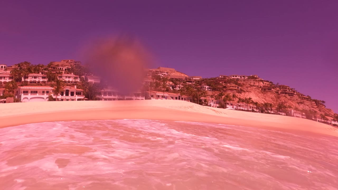 San Jose del Cabo beach view with pink surf in the foreground, pale sand, coastal buildings, palm trees, rocky hills, and a purple-tinted sky, with no recognizable people.