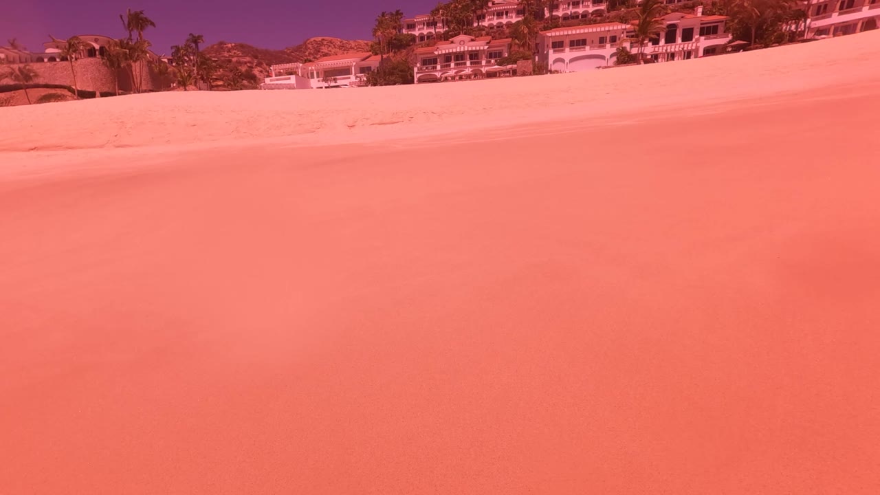 Wide sandy beach in San Jose Del Cabo with hillside resort buildings along the top of frame, pink-orange tint, and no visible people.