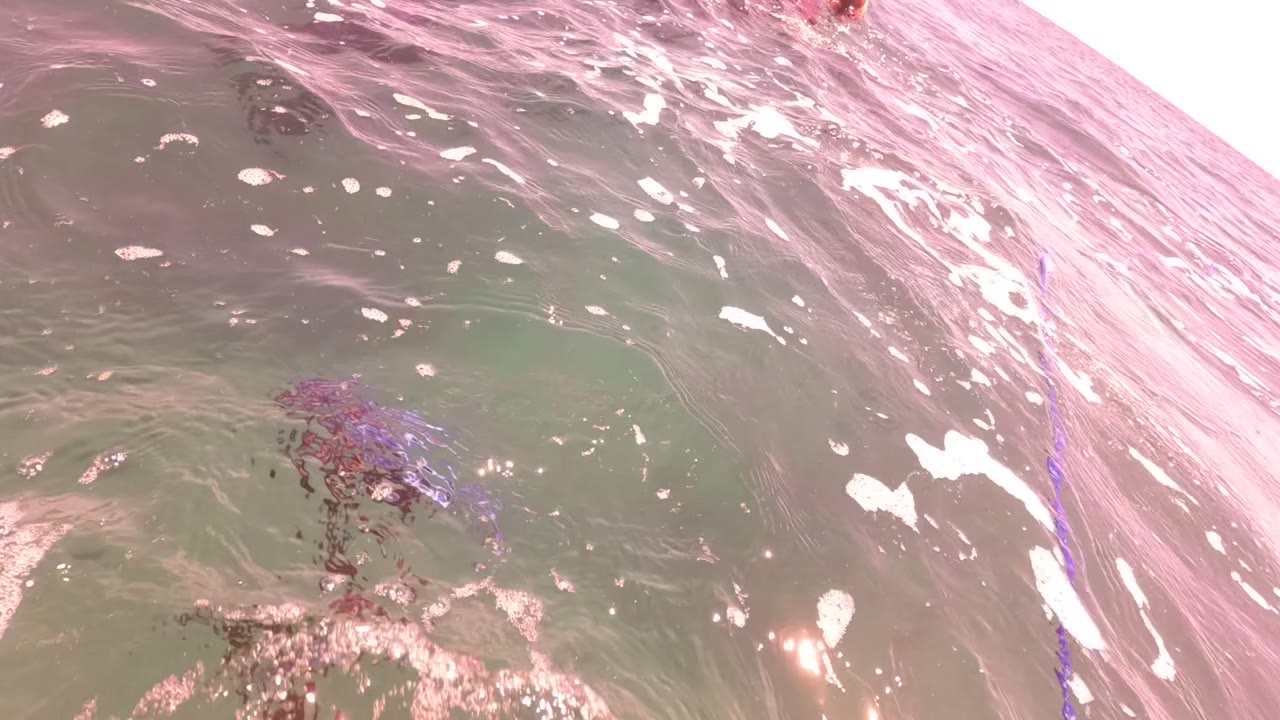 Pink-tinted surface water and foam fill the frame in San Jose del Cabo, with a purple kite line crossing near the right side and no visible people.