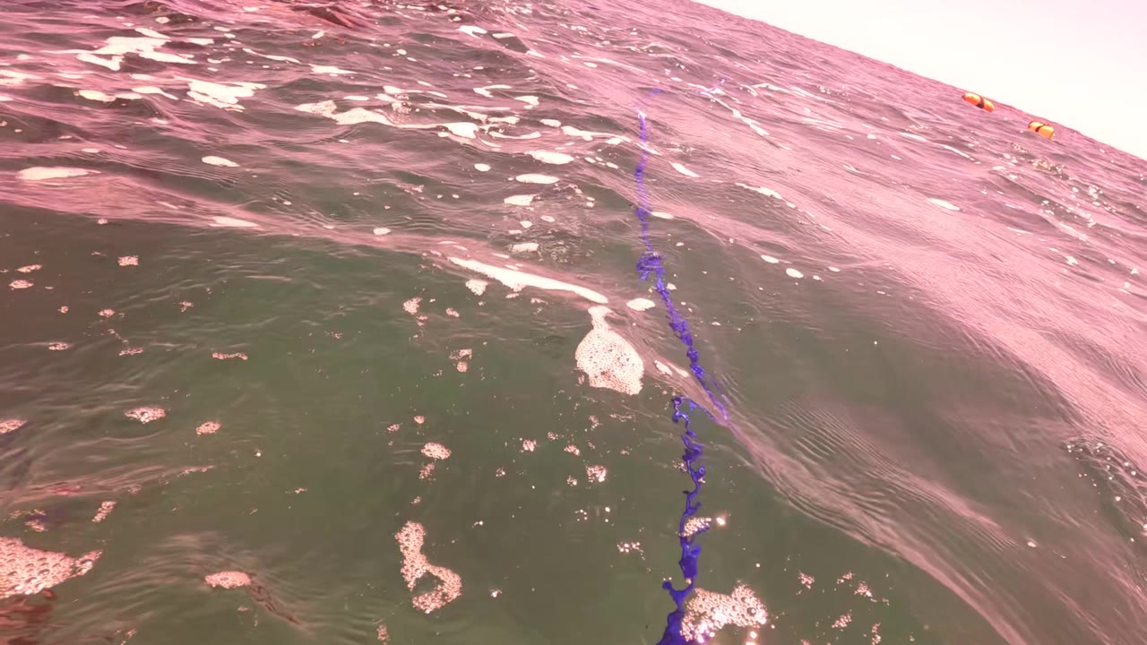 A low waterline view in San Jose del Cabo shows choppy green and pink ocean surface, a vertical purple kite line, foam patches, and no visible people.