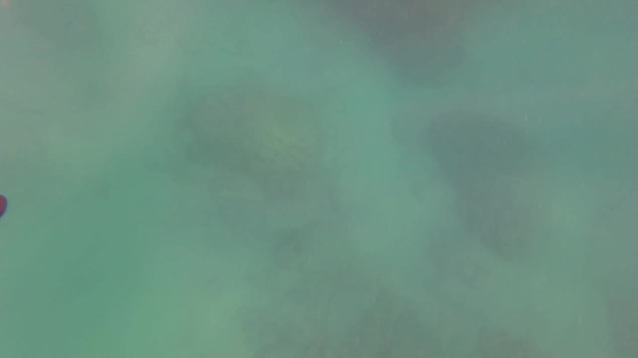 Clear turquoise water in San Jose Del Cabo shows muted rocks below and a small red object near the upper midframe, with no visible people.