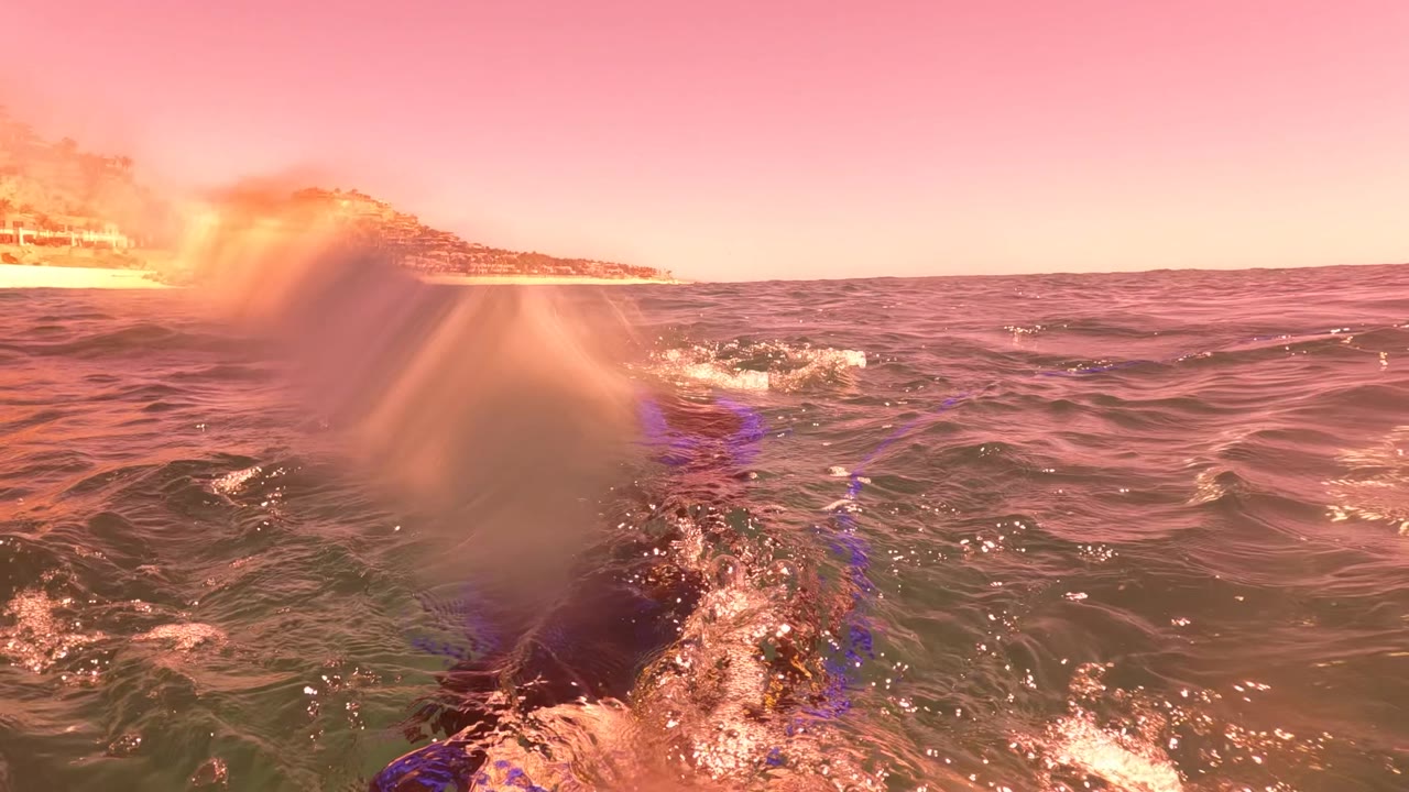 At San Jose Del Cabo, ocean waves fill the foreground as a splash covers part of the lens under pink sunset light, with distant coastline visible.