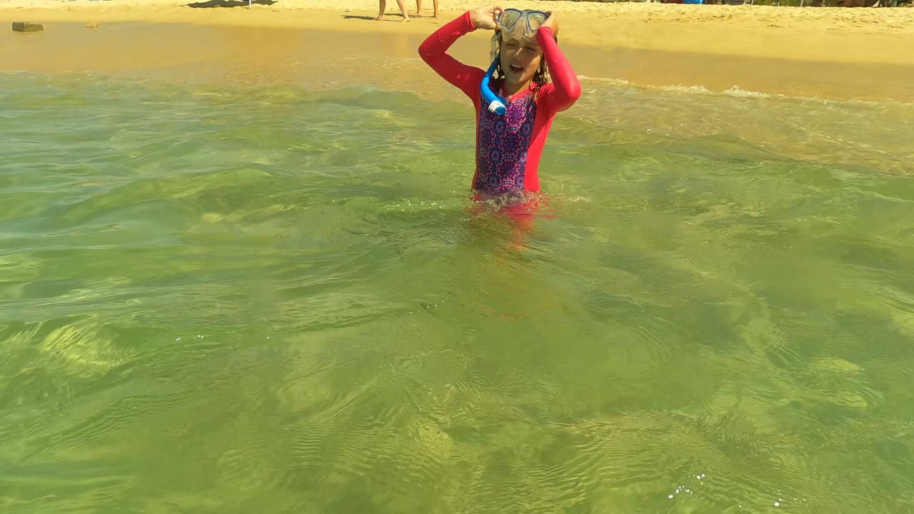 A child in San Jose del Cabo stands chest-deep in green shallow surf, holding a snorkel mask against their face with sandy beach behind.