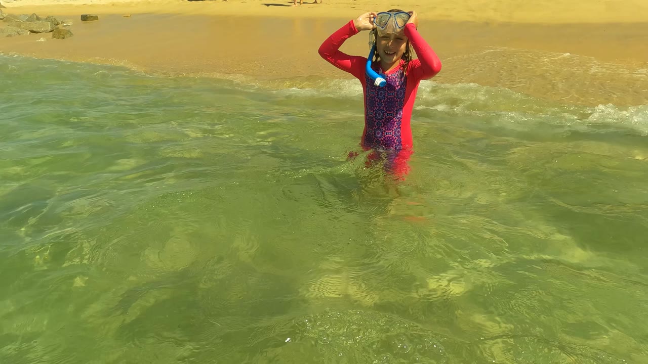Child in red swim shirt holds snorkel mask in shallow surf.