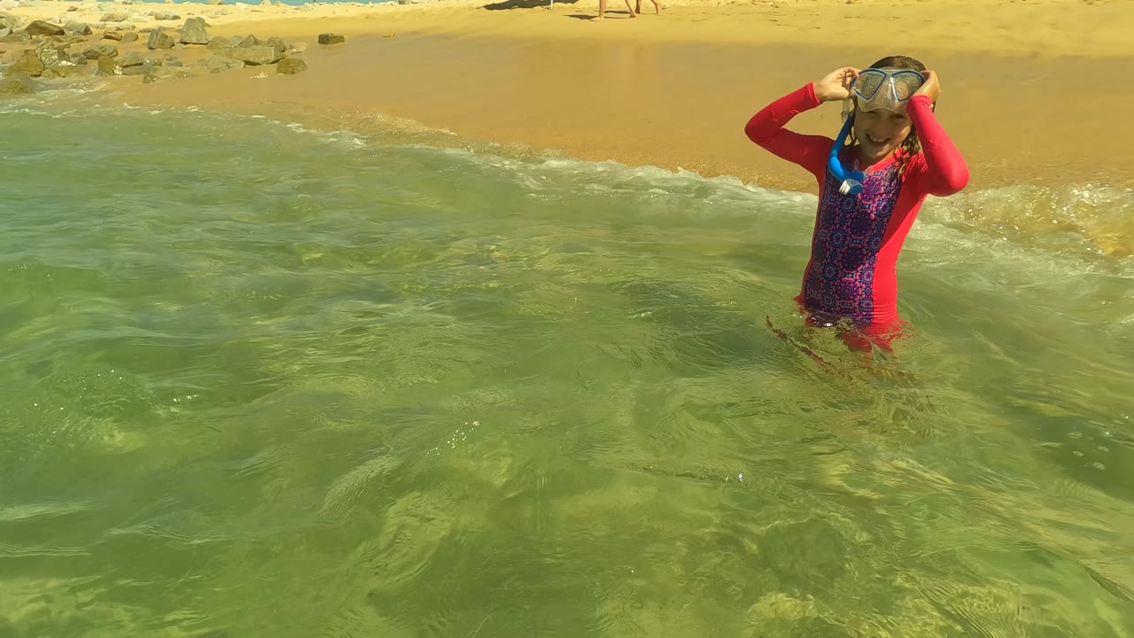 The child appears at the right side of the San Jose del Cabo shoreline frame, still touching the snorkel mask while small waves break behind them.