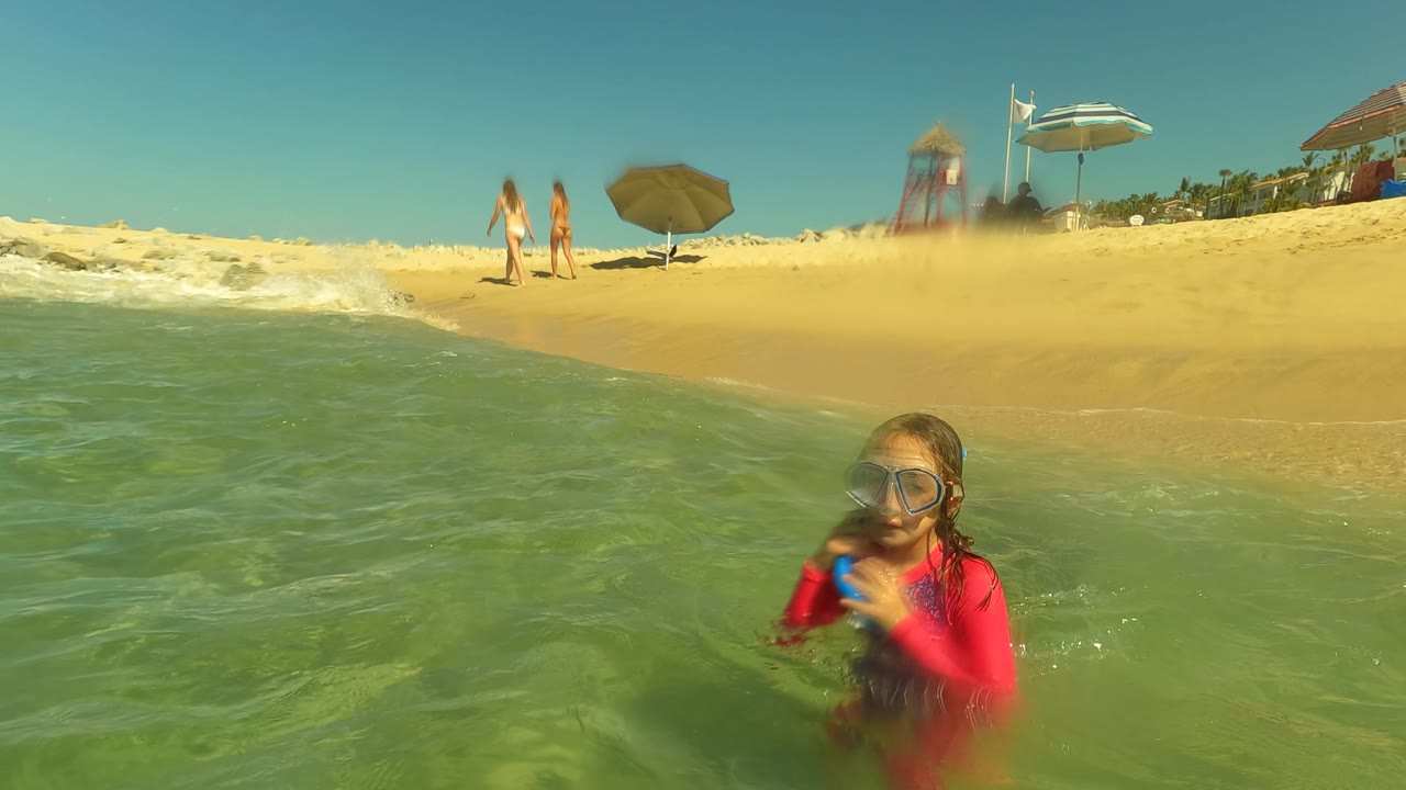 A child wearing goggles and a red swim shirt stands in shallow green-blue water near the sandy beach at San Jose Del Cabo, with umbrellas, walkers, and a lifeguard tower in the background.