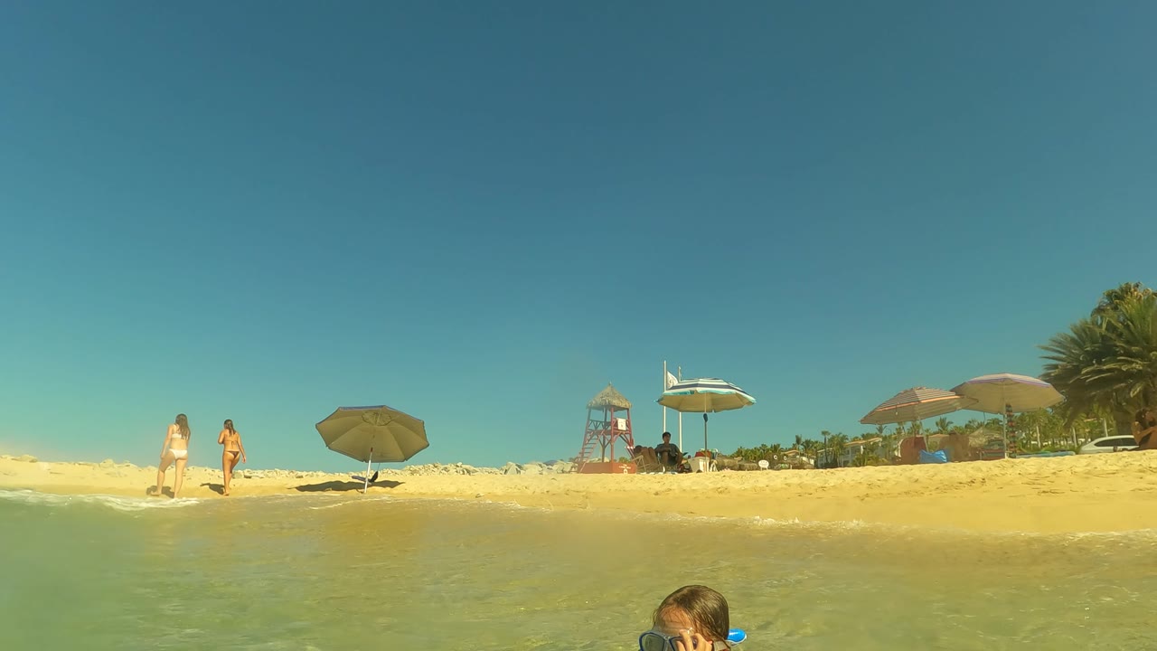 The child is partly cropped at the bottom edge while the San Jose Del Cabo shoreline shows beach umbrellas, people walking on sand, a lifeguard tower, and clear blue sky.