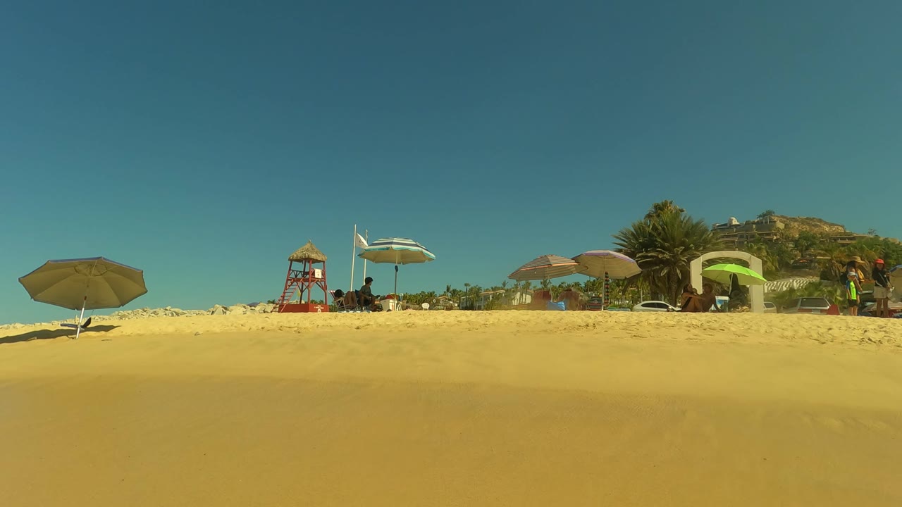 San Jose del Cabo beach view with golden sand in the foreground, umbrellas and a red lifeguard tower along the upper beach, and small beachgoer figures visible in the distance.