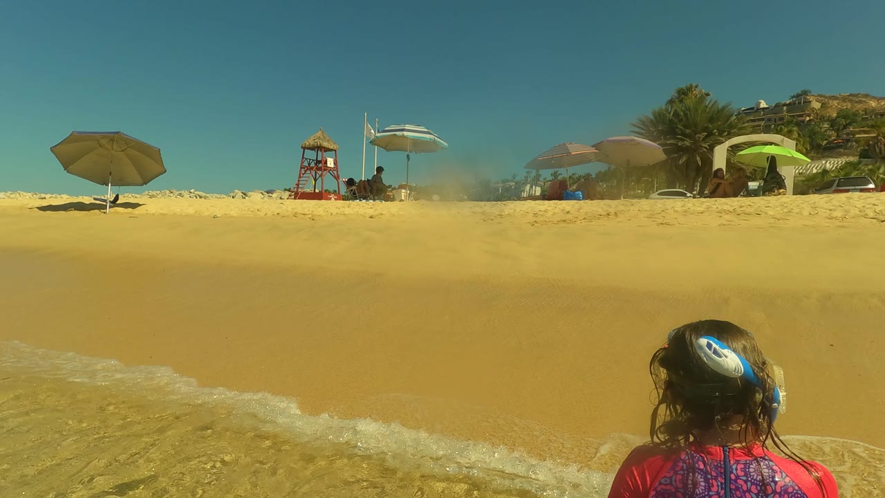 Low beach view with surf foam, umbrellas, lifeguard tower, and people on the sand.