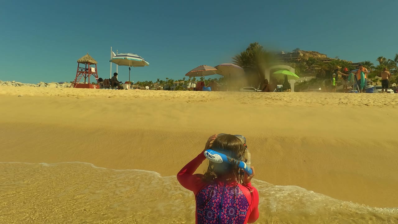 A child in a red shirt with blue hair ties faces the waves at a San Jose del Cabo beach, with foam at the lower edge and umbrellas, people, a palm tree, and a lifeguard tower behind the sand slope.