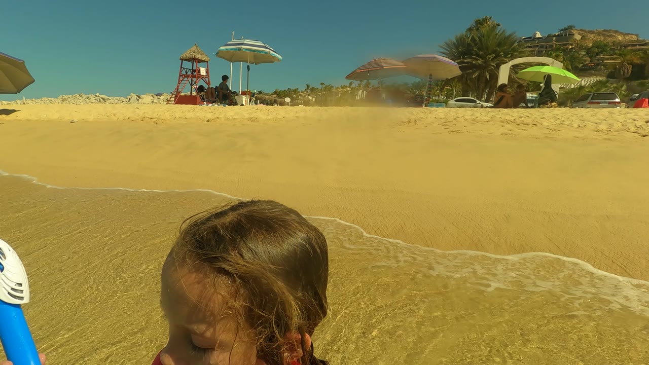 At 0:00 in San Jose del Cabo, a child appears close to the lower-left foreground while a thin wave line crosses wet sand below beach umbrellas and people in the background.